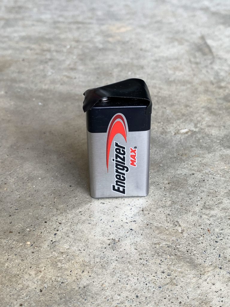 A slightly damaged 9-volt Energizer Max battery with a bent and blackened top rests upright on a concrete surface.
