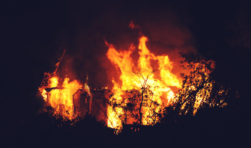 A large house engulfed in bright orange flames at night, surrounded by silhouetted trees. The intense fire illuminates the dark sky, creating a stark contrast with the surroundings.