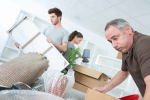 A man in a brown shirt is concentrating on packing a box. Nearby, a young man holds a picture frame, while a woman with a ponytail wraps a lamp. The scene involves moving or packing items, with boxes and packing materials visible.