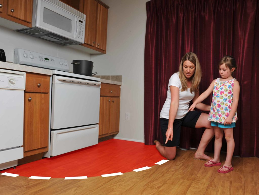 A woman kneels on a kitchen floor with a young girl, pointing to a red semi-circle on the floor in front of an oven and stove. The woman appears to be explaining something about the area marked in red.