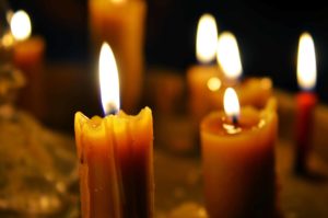 A cluster of lit candles with melting wax casting a warm, soft glow in dim lighting. The flames flicker gently, creating a serene and peaceful atmosphere. The background is dark, highlighting the candlelight.