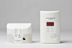 Two white carbon monoxide alarms are shown. The left one is compact with a button labeled "Test/Reset." The right one is taller with a digital display reading "8.8.8" and buttons marked "Test/Reset" and "Peak Level.