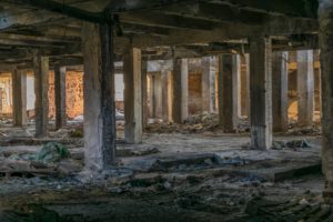 Abandoned building interior with exposed concrete pillars and debris scattered across the floor. Walls are partially missing, revealing some brickwork. Sunlight filters through openings, creating a pattern of light and shadow on the ground.