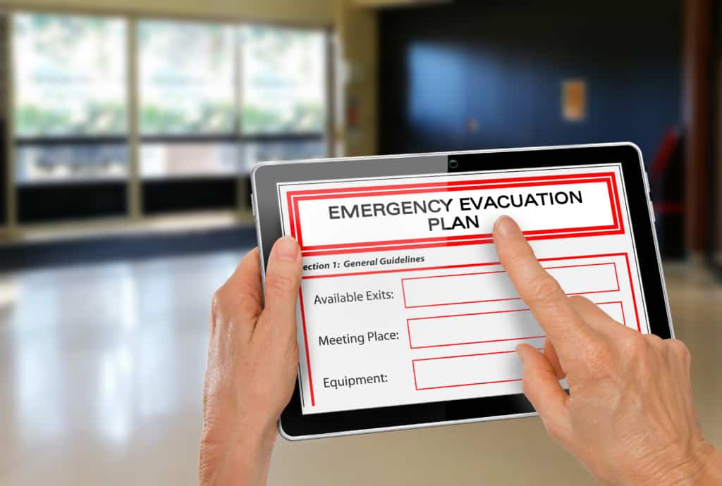 A person holds a tablet displaying an "Emergency Evacuation Plan" with sections for available exits, meeting place, and equipment. The background shows a blurred indoor space with large windows.