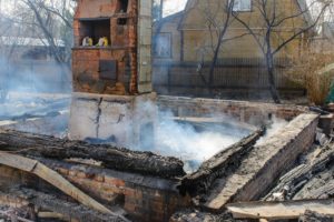 Burned-down house with only the brick chimney and foundation remaining. Smoke rises from charred wooden beams and rubble scattered around. In the background, a wooden fence and leafless trees are visible.
