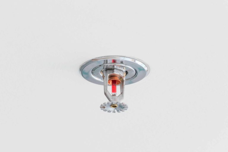 A ceiling-mounted fire sprinkler with a metallic finish is centered against a plain white background. The sprinkler has a red glass bulb inside, indicating its activation mechanism.
