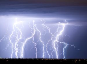 A night sky is dramatically lit up by multiple bright, jagged lightning bolts striking down. Below the storm, distant city lights are visible on the horizon beneath a dark, heavy cloud cover.