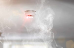 Smoke surrounds an activated ceiling-mounted smoke detector with a glowing red light, set against a blurred background.