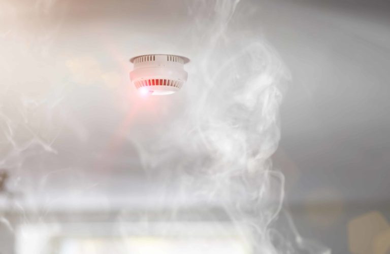 Smoke surrounds an activated ceiling-mounted smoke detector with a glowing red light, set against a blurred background.