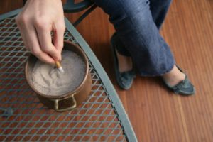 A person in jeans and moccasins stirs a drink in a brown mug with a spoon on a metal mesh table. The background shows a wooden floor.