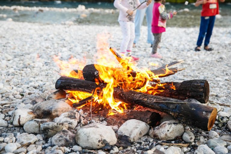 A campfire burns brightly on a stony riverbank, surrounded by large rocks. In the background, children stand near the water's edge, partially blurred, enjoying the outdoor setting.