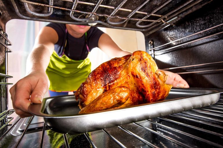 A person wearing a green apron places a roasted chicken into an oven using a metal baking tray. The chicken is golden-brown and coated with a crispy skin. The perspective is from inside the oven looking outward.
