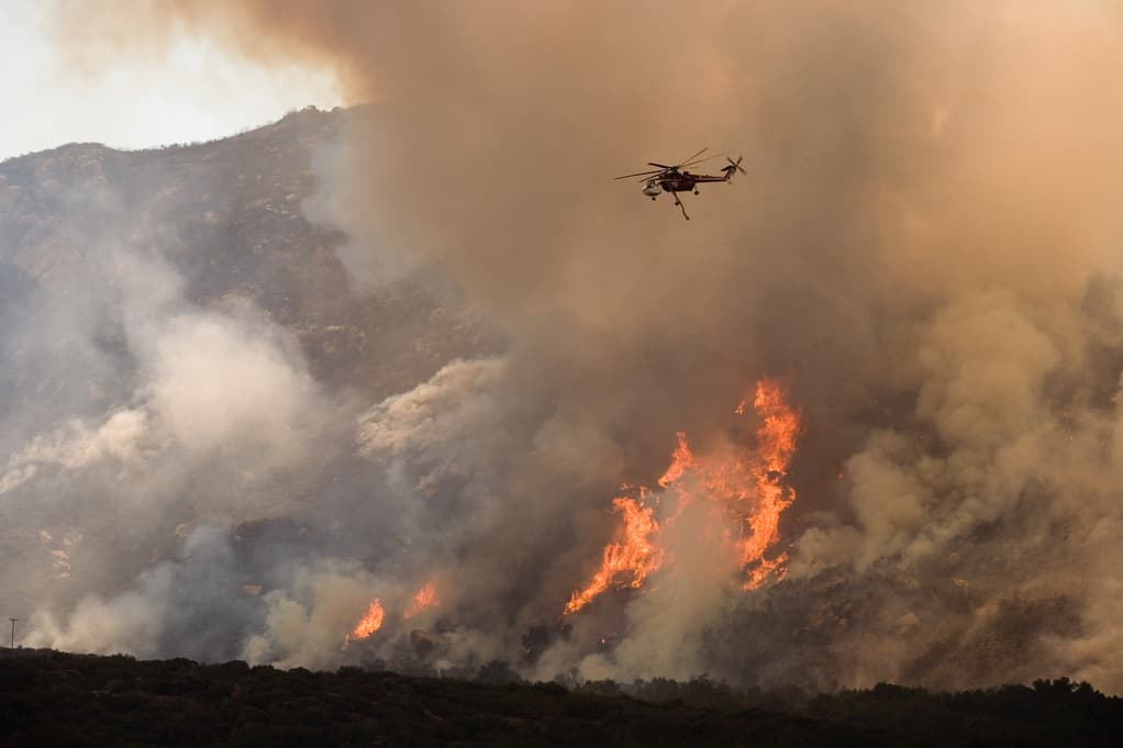 A helicopter flies above a large wildfire on a mountainside, dropping water to combat the spreading flames. Thick smoke billows into the sky, contrasting with the bright orange and red of the fire. Dense vegetation is visible in the foreground.