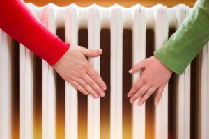 Two hands wearing long-sleeve shirts, one red and one green, are held close to a white radiator. The position suggests the individuals are feeling the warmth emanating from the heater.