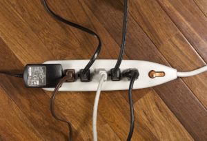 A power strip with several black, brown, and white cords plugged in is lying on a wooden floor. The cords are connected to various devices, with one unplugged socket visible.