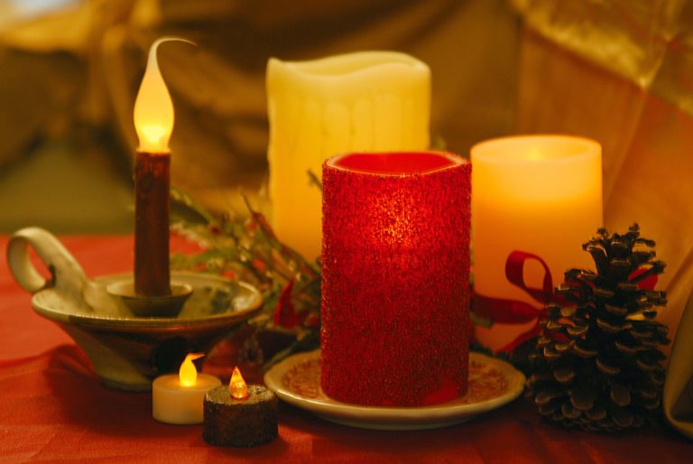 A cozy candle arrangement on a red tablecloth features a mix of glowing flameless candles and a single wax candle. A glittery red candle stands out alongside pine cones and festive greenery, creating a warm, inviting atmosphere.