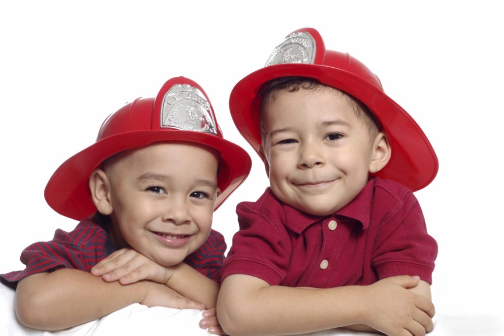 Two young boys smiling and wearing red toy firefighter helmets. They are leaning on their folded arms and wearing red shirts. The background is plain white.