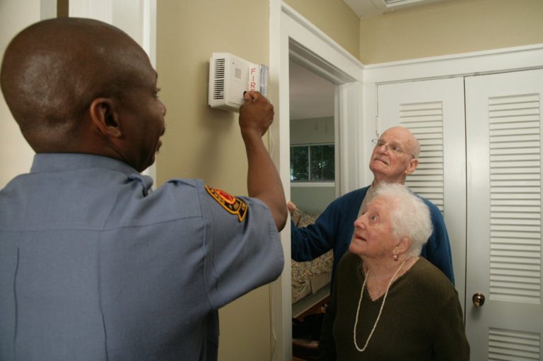 A firefighter in uniform demonstrates a fire alarm to an elderly couple in a home. The man points to the alarm, while the couple attentively observes. They are standing in a hallway near a door.