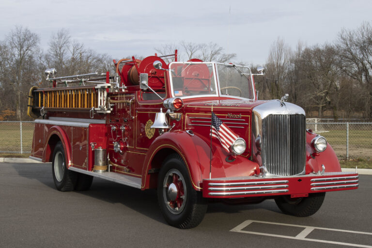 A vintage red fire truck with chrome details and an American flag on the front, parked on a road with leafless trees in the background. The truck features ladders and hoses, showcasing a classic design.