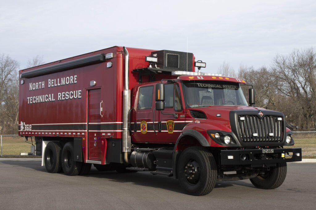 A red technical rescue truck with "North Bellmore Technical Rescue" written on the side is parked on a paved area. The vehicle has an extended cargo area and the surroundings include bare trees and a chain-link fence.