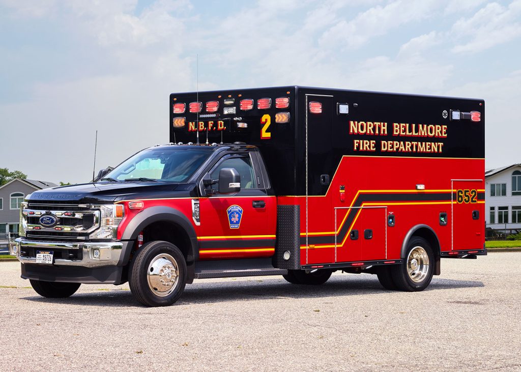 A red and black North Bellmore Fire Department ambulance is parked on a paved surface under a cloudy sky. It has visible emergency lights and the number 2 on the side. Buildings are faintly visible in the background.