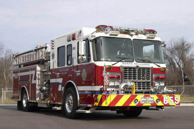 A red and white fire truck is parked on a paved surface. It features multiple lights and equipment compartments. The front has bold numbering "655" and American flag details. Trees and a fence are visible in the background.