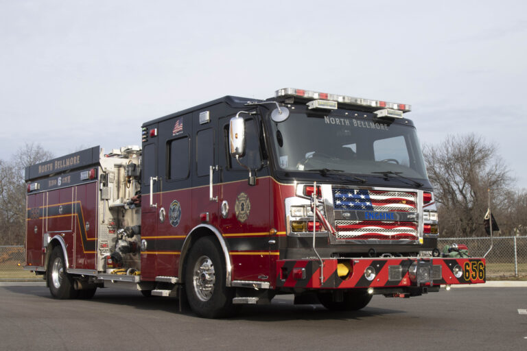 A red and black fire truck with the words "North Bellmore" on it, parked on a paved road. The front grille features an American flag design, and the truck has various firefighting equipment and reflective markings. Trees and a fence are in the background.