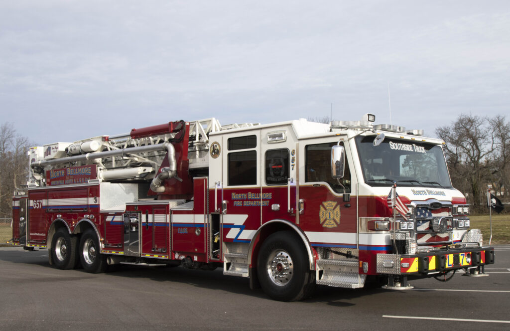 A red and white fire truck with various equipment and ladders is parked on a paved road. The vehicle is marked with "North Bellmore Fire Department" on the side. The background has trees and a grassy area under a cloudy sky.