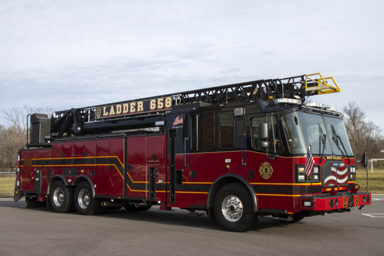 A vibrant red fire truck with a tall, extended ladder on top, labeled "Ladder 658" on the side. The vehicle is stationary on a paved area with a grassy field and trees in the background under a cloudy sky.