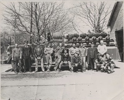 A vintage black and white photo of a group of firefighters posing in front of a fire truck. The men are wearing uniforms and casual attire. Leafless trees and a building are in the background.