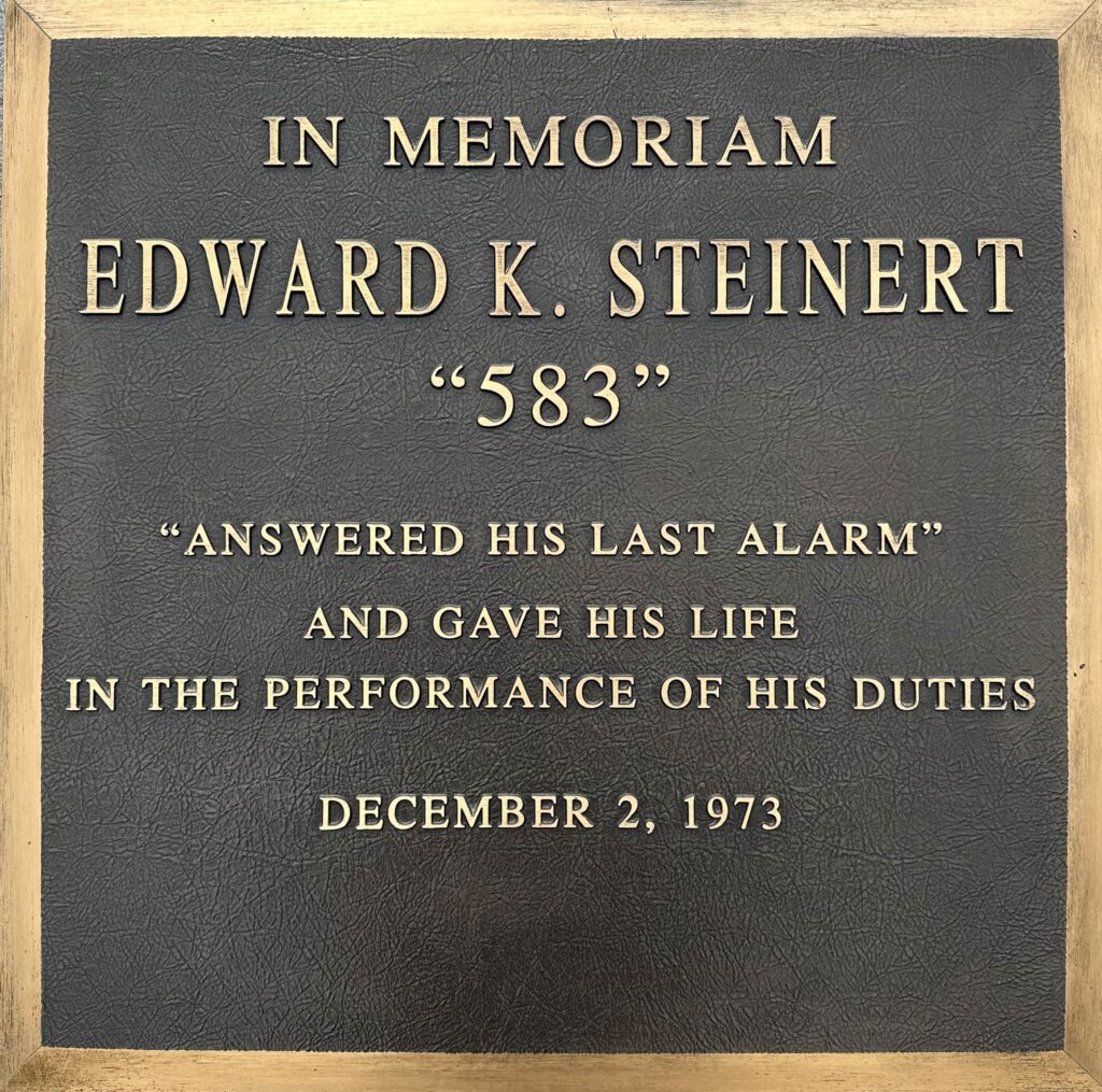 Plaque with the inscription: "In Memoriam Edward K. Steinert '583'. 'Answered his last alarm' and gave his life in the performance of his duties. December 2, 1973." The plaque is set in a wooden frame.