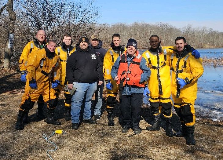 A group of nine people, including eight wearing yellow protective suits and one in a red vest, pose outdoors by a lake. The background features trees and a partly cloudy sky. The ground is grassy and scattered with patches of ice.