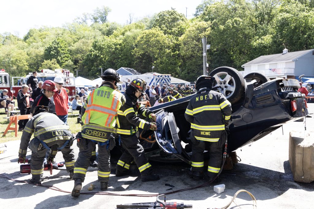 Firefighters in full gear work to rescue someone from an overturned car during a public demonstration, with onlookers and emergency vehicles visible in the background.